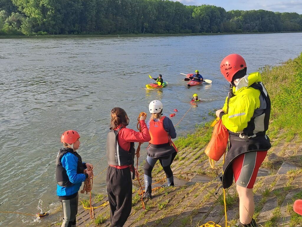 Foto: Vom Ufer aus retten Kanuten eine gekenterte Person mittels Wurfsack und Rettungsleine. Auf dem Wasser sichern zwei Paddler die Gefahrensituation und schleppen das havarierte Kajak mit einem Cowtail-Rettungsseil an Land (I. Gohle).